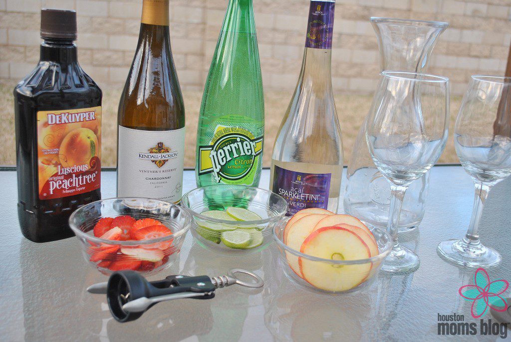 Ingredients for white sangria including peach schnapps, white wine, sparkling water, sparkling wine, strawberries, limes, apples, wine glasses, a pitcher, and a wine opener. Logo: Houston moms blog. 