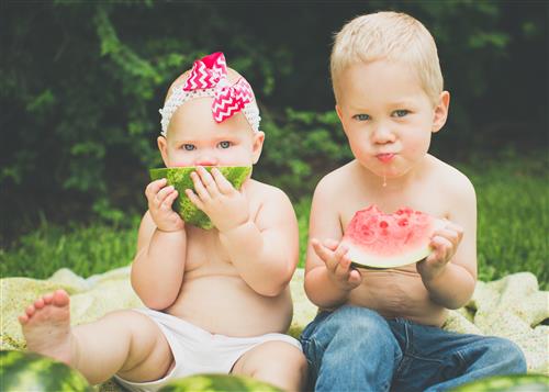 baby and toddler sit on blanket in grass eating watermelon