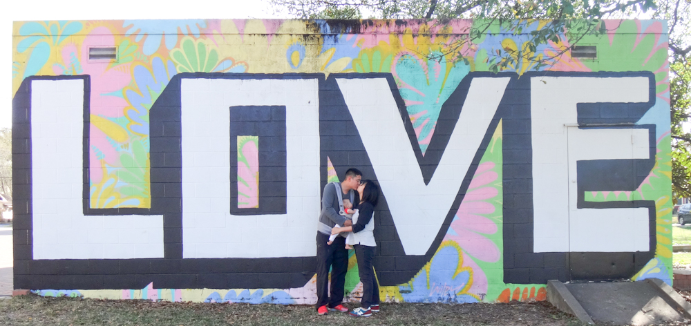 Two people holding a baby and kissing in front of a mural with the text: Love. 