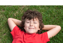 A photograph of a smiling child lying on the grass and wearing a red shirt.