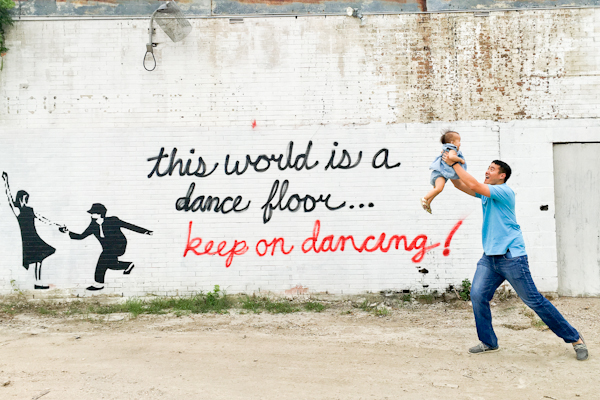 A man holding a baby in the air in front of a mural which reads this world is a dance floor... keep on dancing. 