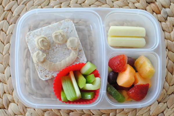 A plastic container with three divisions containing a fruit salad, a cheese stick, a rice cake with hummus drawn on it in the form of a smiley face and a muffin cup of celery sticks. 