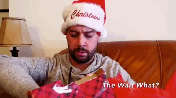 A GIF of a confused man wearing a Santa hat opening a present and pulling out a simple painting of flowers. Text states: The wait what?