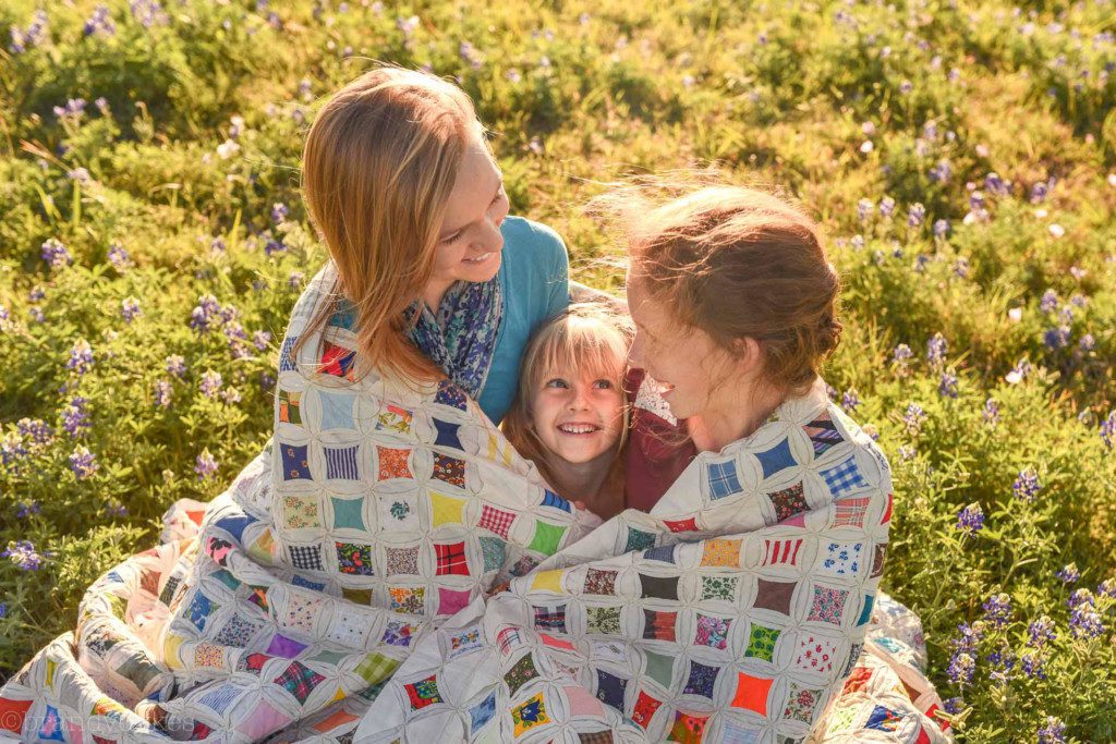 A smiling mother and two children wrapped in a quilt and sitting in a field of bluebonnets. 