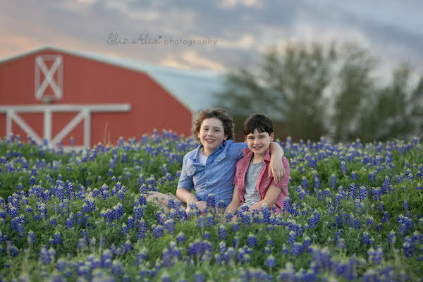 Two children with their arms around each other sitting in a field of Bluebonnets. A barn is in the background. 