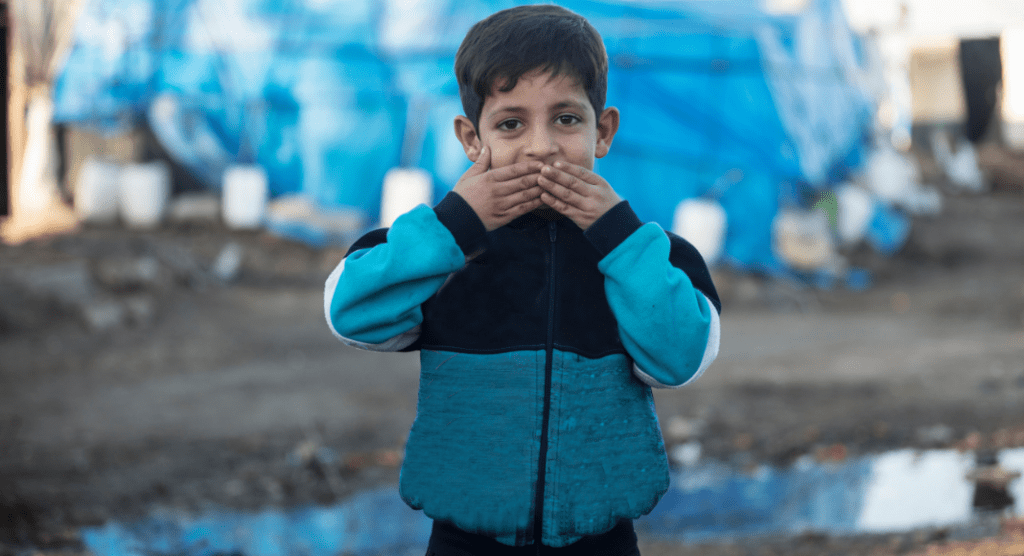 A refugee child pressing his hands to his lips. 