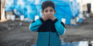 A refugee child pressing his hands to his lips.