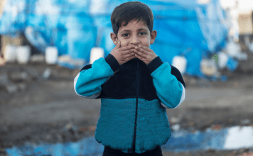 A refugee child pressing his hands to his lips.