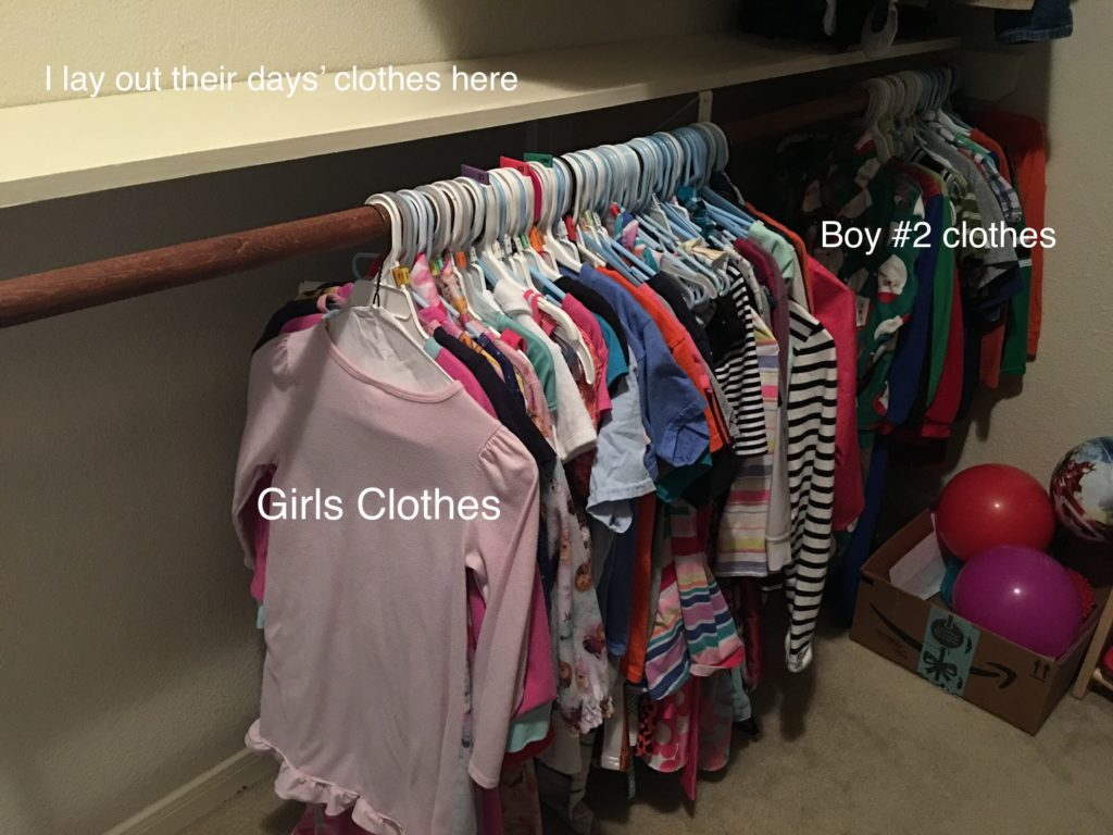 A closet. A shelf is labeled I lay out their days' clothes here. Beneath the shelf are two sets of hanging clothes labeled Girls clothes and boy number 2 clothes. 