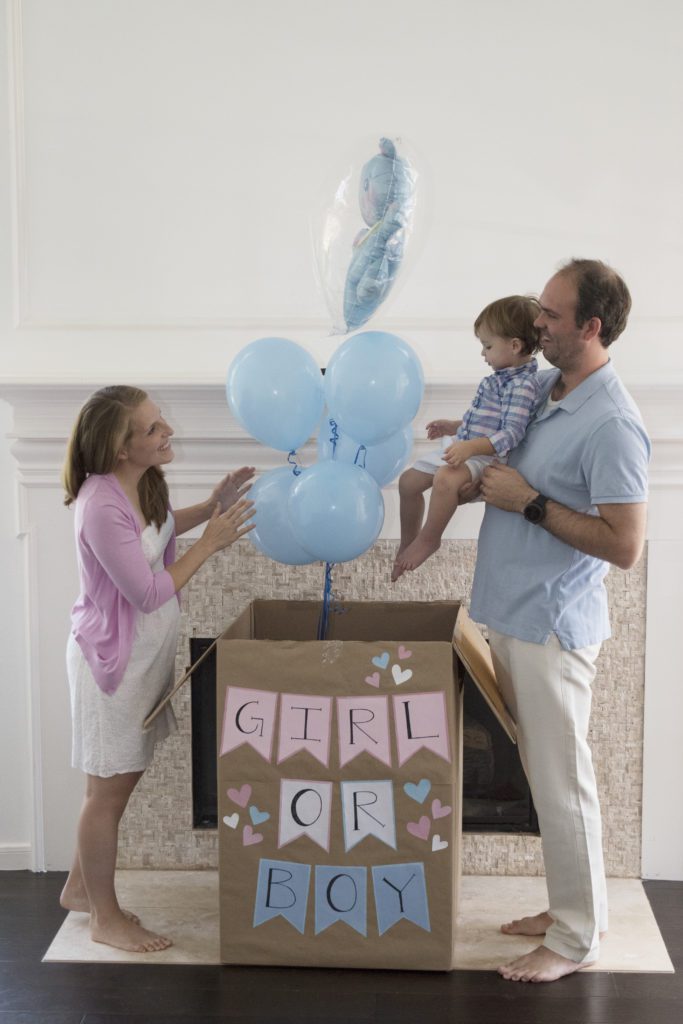 A mother and a father holding a young child stand next to a box labeled girl or boy. Extending out of the opened box are blue balloons. 