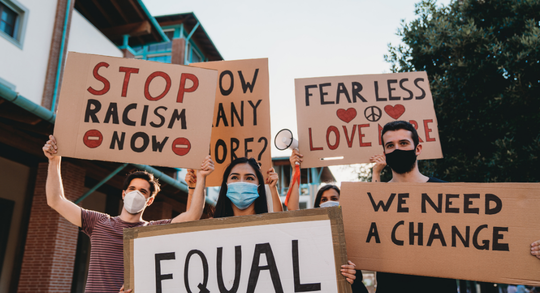 A group of People holding signs with the text: Stop Racism Now. Fear Less Love more. We need a change.