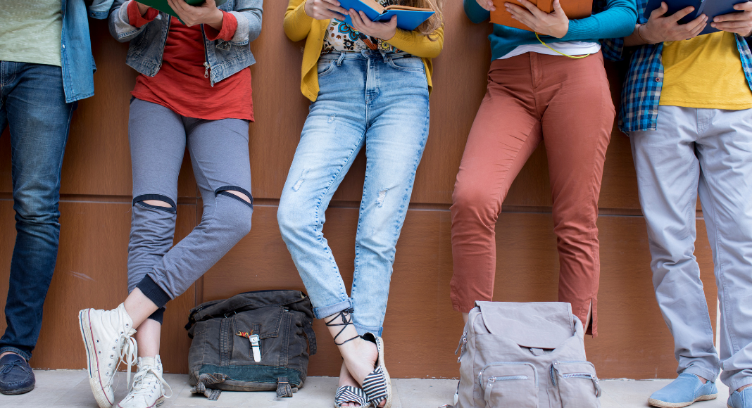 A photograph of five people wearing jeans and holding open books with backpacks at their feet.