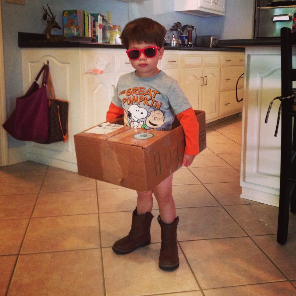 A photograph of a young child wearing cowboy boots and sunglasses and with a box with a hole cut out of it around his waist. 