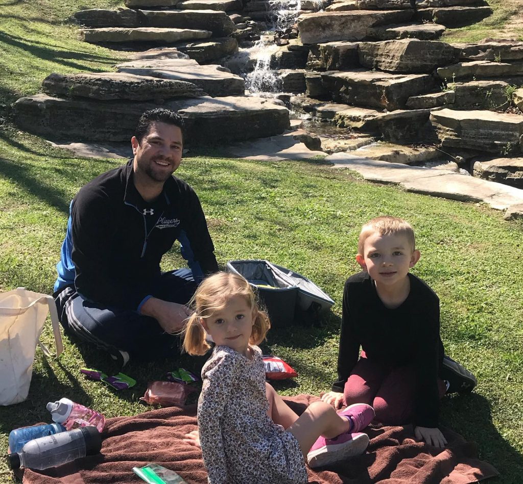 A father and two children sitting on top of a picnic blanket on the grass with a waterfall in the background