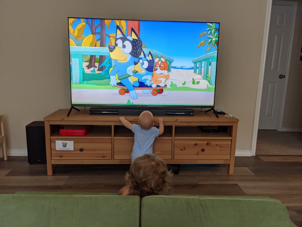 A baby and a young child watching Bluey on a TV at home. 