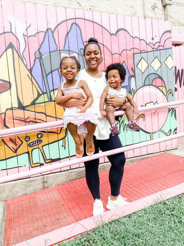 A mother holding two young children while standing in front of a mural. 