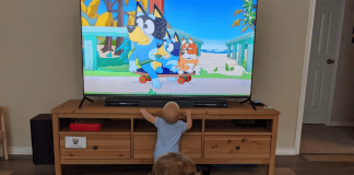 A baby and a young child watching Bluey on a TV at home.