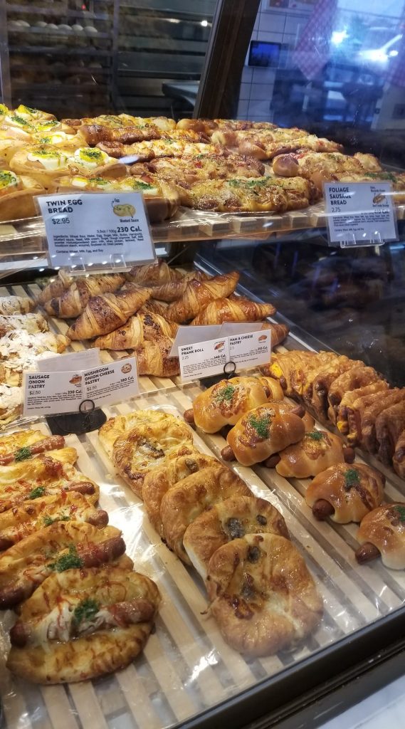 A store glass case with different types of pastries including twin egg bread, foliage sausage bread and mushroom, onion, cheese pastry. 