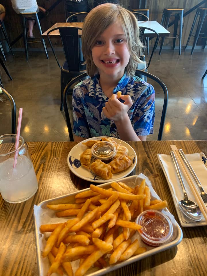 A smiling child sitting at a table in a restaurant with Asian dumplings and fries on the table. 