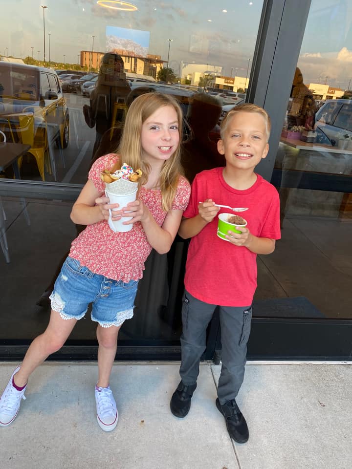 Two smiling children holding food containers and standing in front of a restaurant. 
