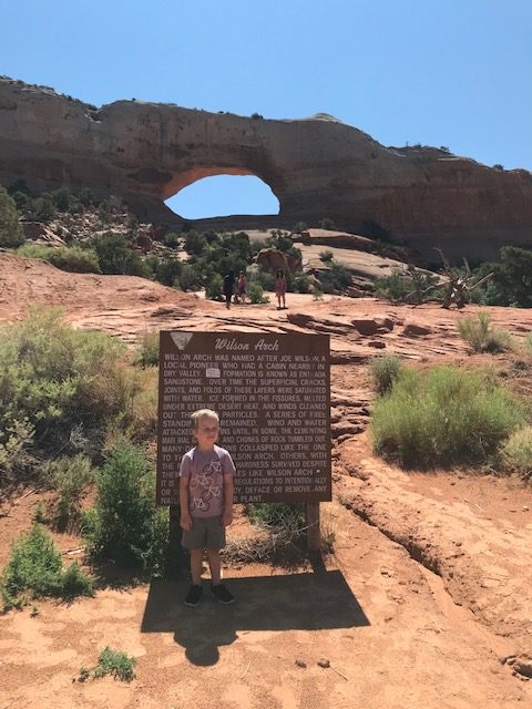 A child standing in front of a sign for Wilson Arch with a rock arch in the background. 