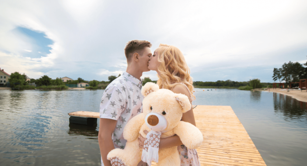 A young woman holding a large teddy bear and kissing a young man's cheek on a dock with a lake in the background. 