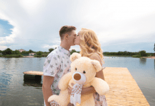 A young woman holding a large teddy bear and kissing a young man's cheek on a dock with a lake in the background.