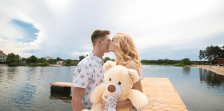 A young woman holding a large teddy bear and kissing a young man's cheek on a dock with a lake in the background.