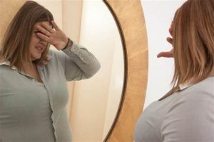 woman covers her eyes as she looks at her body in the mirror