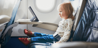 A toddler sits on an airplane seat watching an ipad on the tray table.