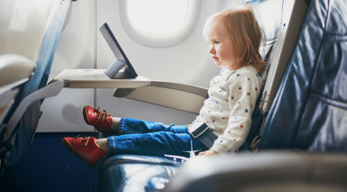 A toddler sits on an airplane seat watching an ipad on the tray table.
