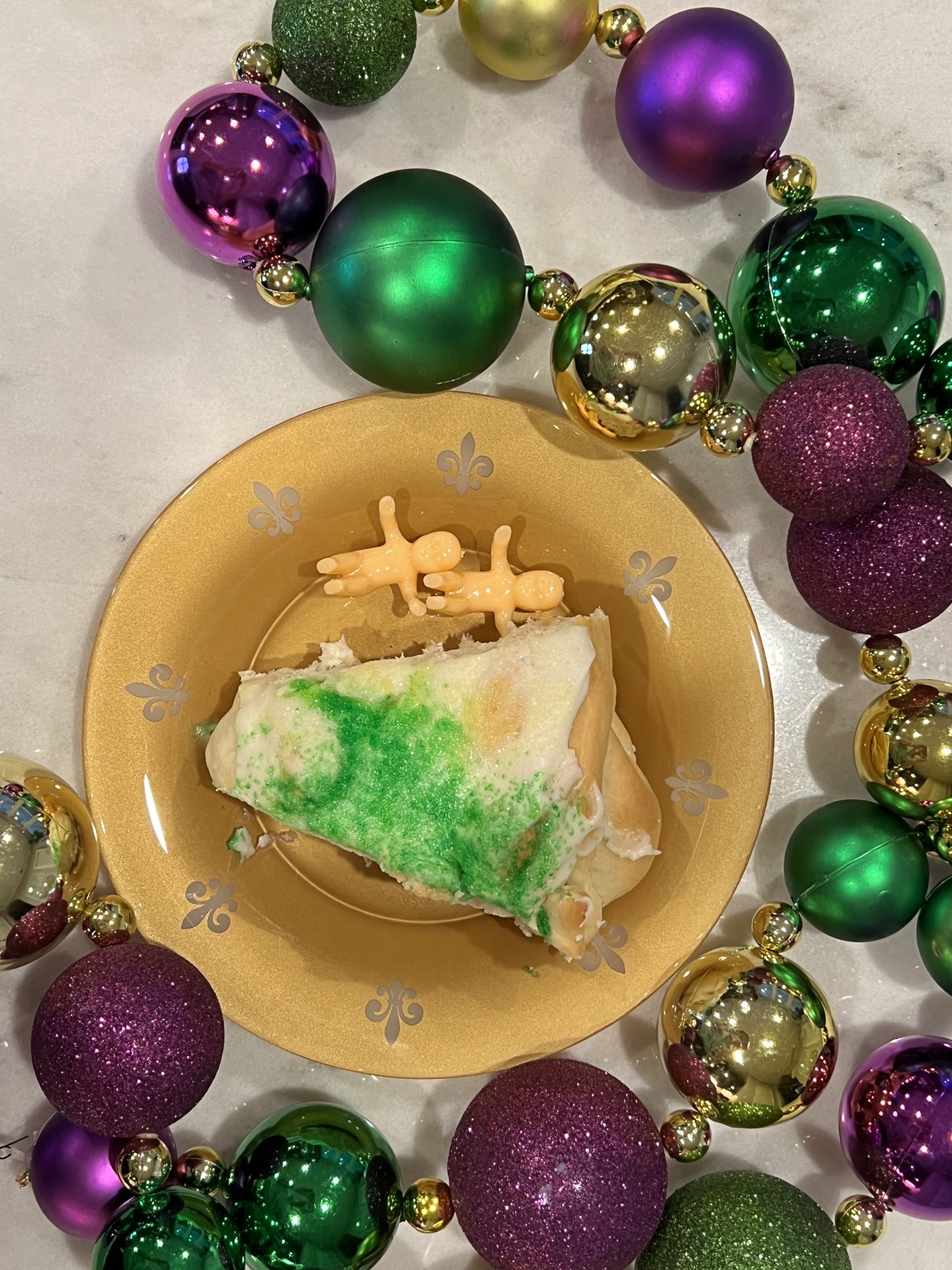 Slice of King Cake with plastic babies on the plate, surrounded by Mardi Gras beads