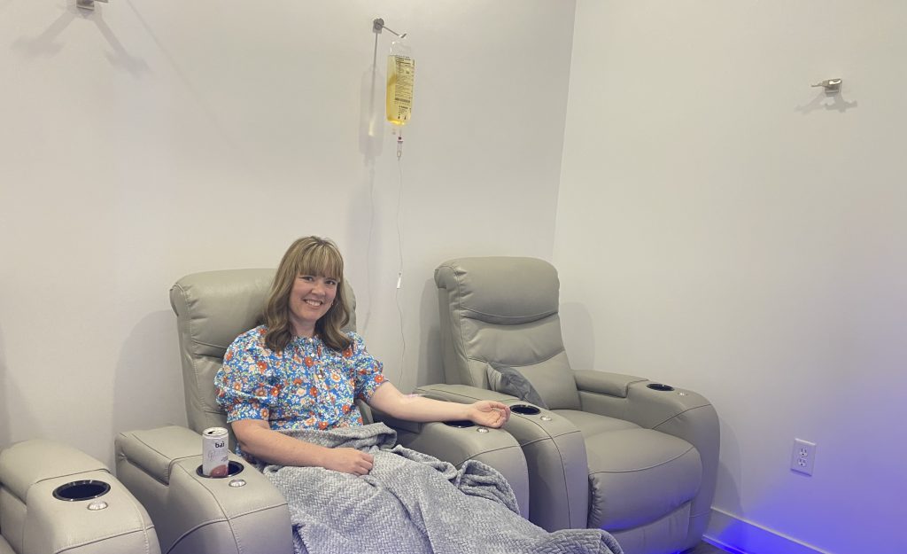woman sits in chair with IV fluid bag hanging all wall behind her