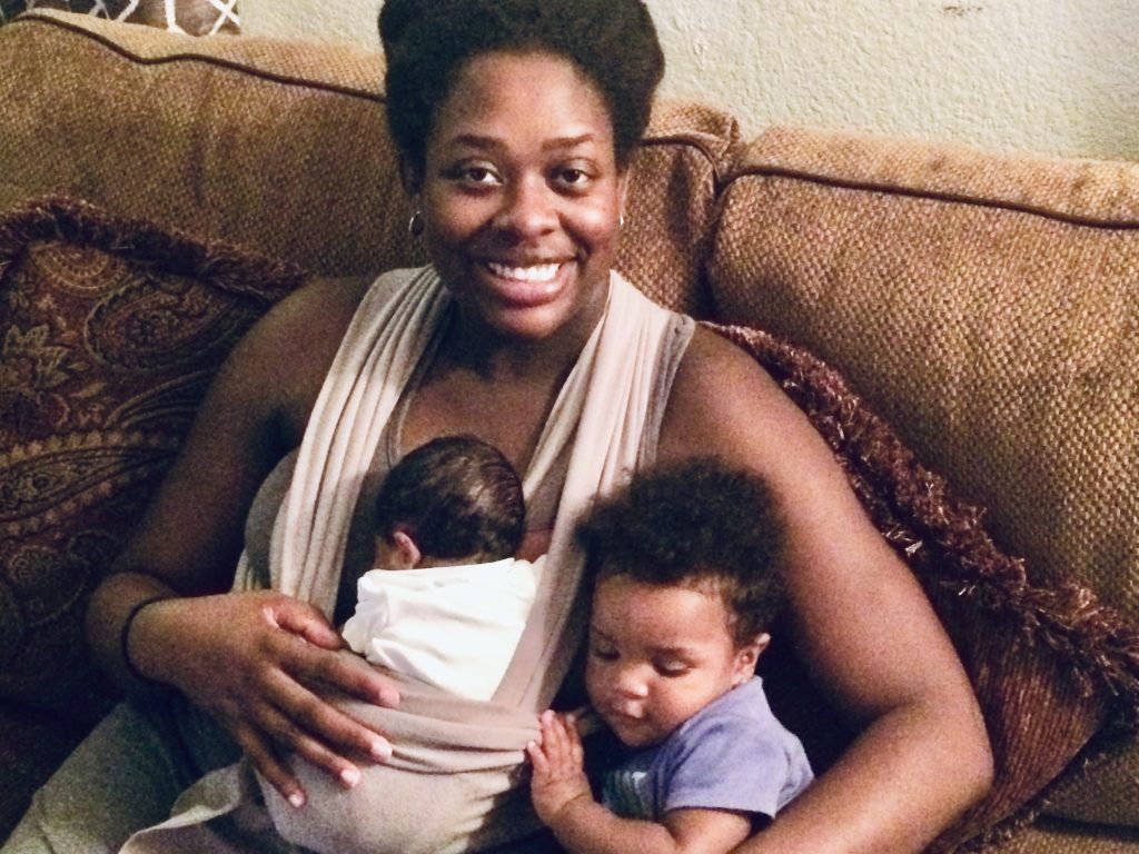 A mother cradles newborn in a sling while cuddling a toddler beside her