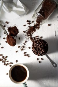 cup of coffee, hard of coffee grounds on its side, and scoop of whole coffee beans on a white table