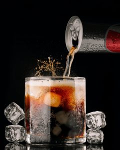 can of soda being poured into a glass with ice cubes beside it