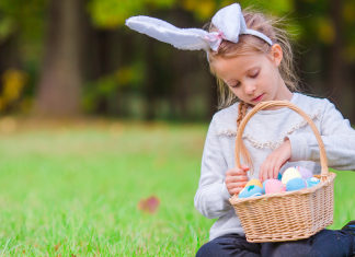 child with bunny ears on sits in grass holding Easter basket full of colored eggs