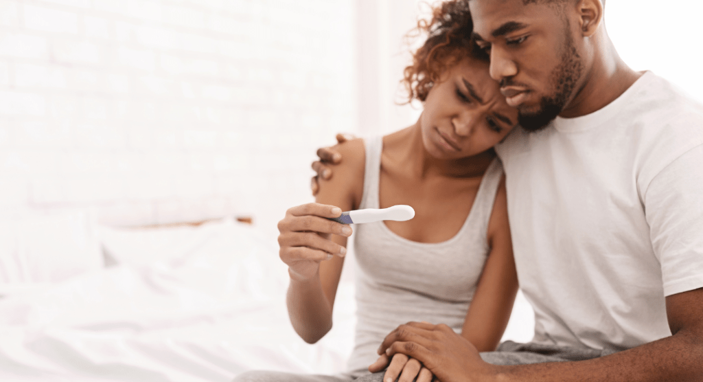 man and woman sit on bed with sad expressions, holding pregnancy test