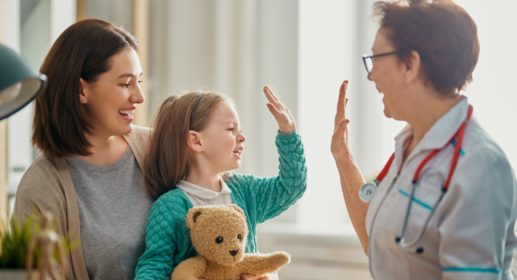medical professional high fives little girl who is sitting in her mother's lap holding a teddy bear