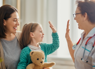 medical professional high fives little girl who is sitting in her mother's lap holding a teddy bear