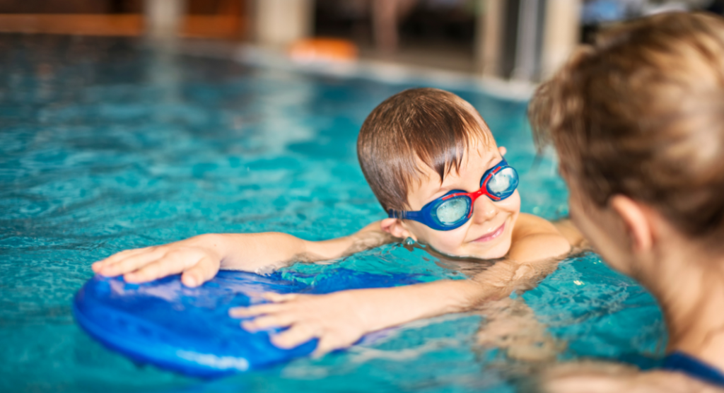 child with goggles looks at swim instructor while holding on to kickboard