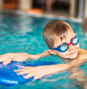 child with goggles looks at swim instructor while holding on to kickboard
