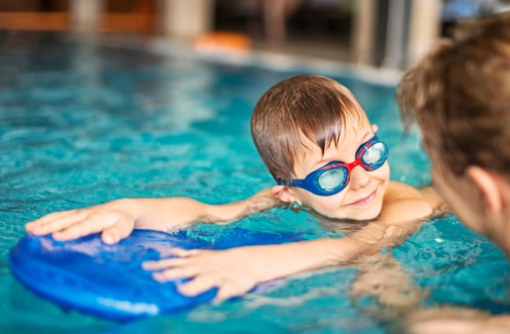 child with goggles looks at swim instructor while holding on to kickboard