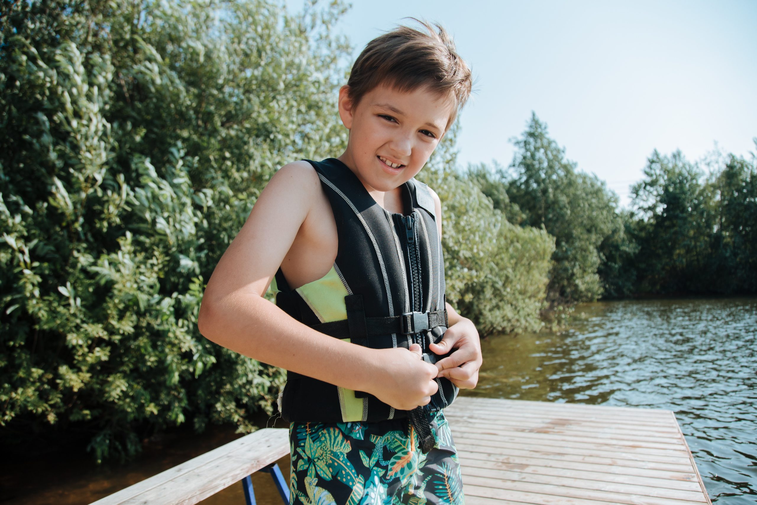 boy wearing life jacket stands on dock of lake