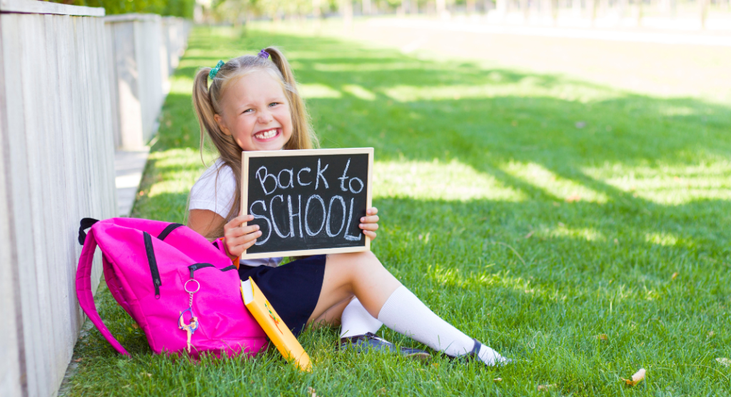 preschool girl sits on grass with Back to School sign and backpack