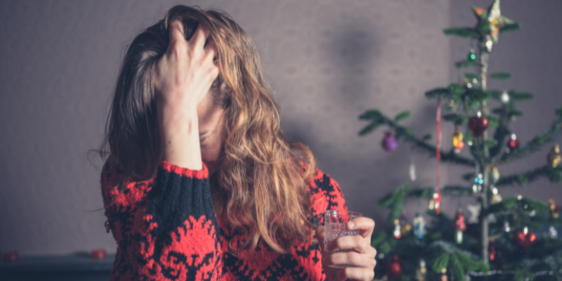 woman in holiday sweater puts head in hand with drink in other hand