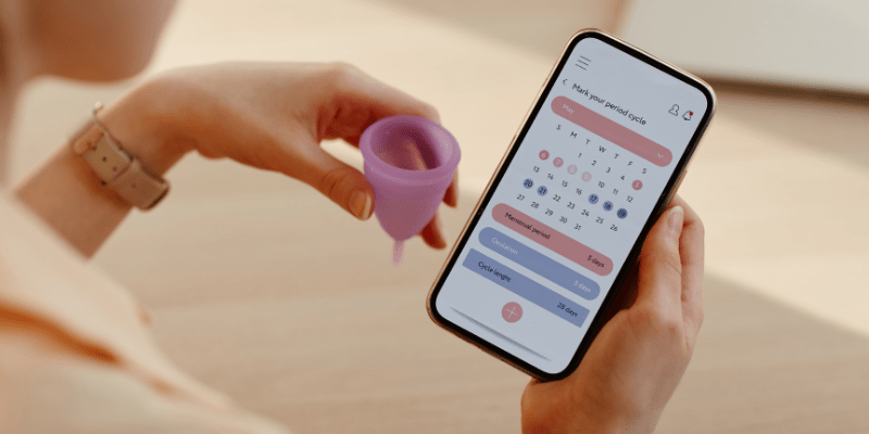 hand holds menstrual cup with phone with cycle tracking app in other hand