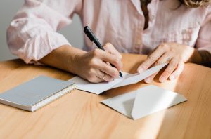 image of woman's hands writing a letter. the woman is wearing a light pink blouse with 3/4 length sleeves. she's holding a black pen, writing in a card with an envelope and notepad nearby on the table