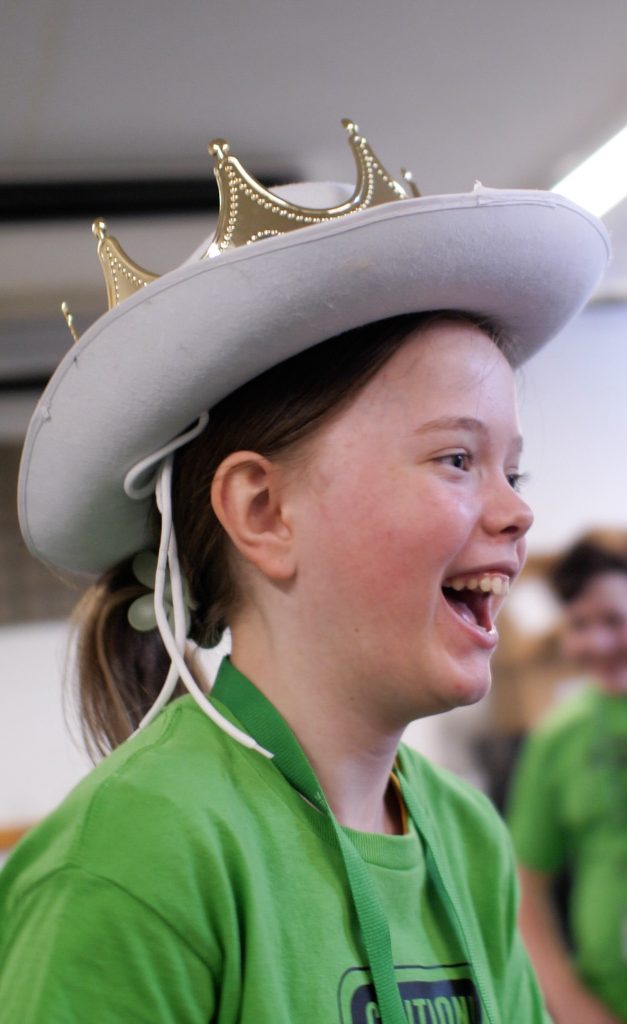 A girl wearing a cowboy hat and a crown laughs at summer camp