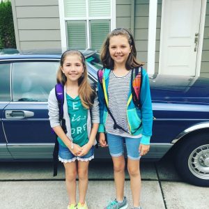 two girls stand in front of a car with backpacks on
